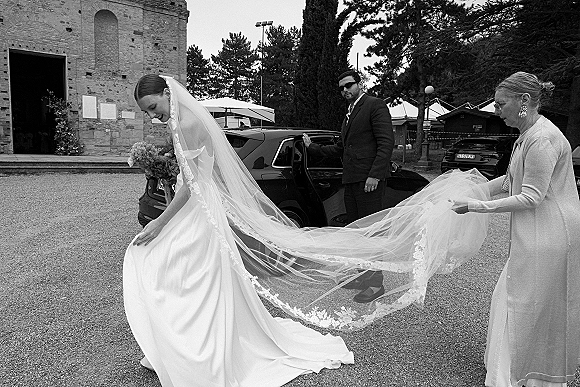 Bride getting ready with long veil and bouquet as family adjusts her lace train in a gravel courtyard by a stone building