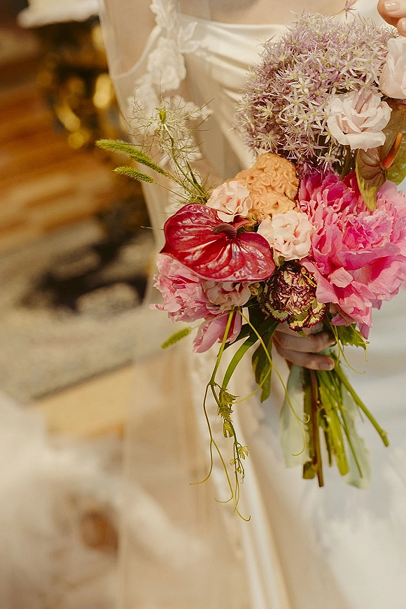 Bridal bouquet with pink peonies and greenery held in the bride’s hand against lace dress detail on stone steps with warm bokeh lights