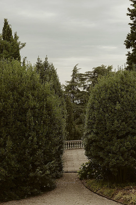 Garden walkway with stone paving and gravel path beside a stone balustrade, bordered by shrubs and trees under a cloudy sky