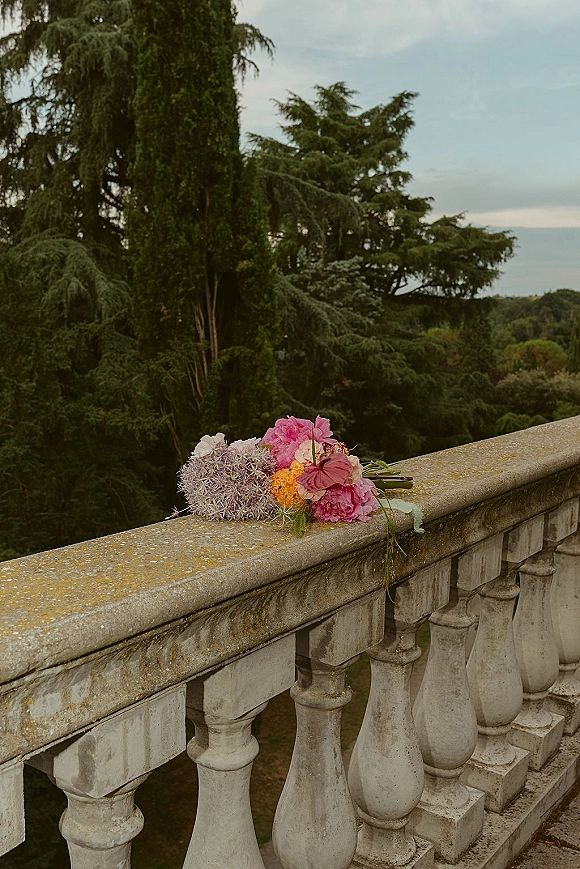 Wedding bouquet with pink peonies and mixed orange blooms, ribbon-wrapped, resting on a stone balustrade overlooking a cloudy garden terrace