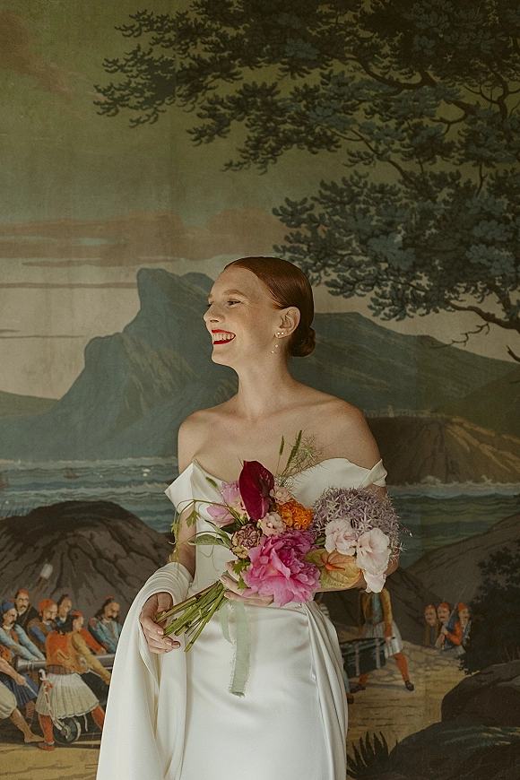 Bridal portrait of bride holding bouquet in an off the shoulder wedding dress with long white gloves and red lipstick before a mural backdrop