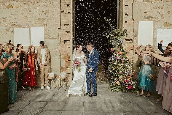 Wedding recessional as bride in satin dress and veil walks with groom in blue suit through confetti under a floral arch in stone courtyard