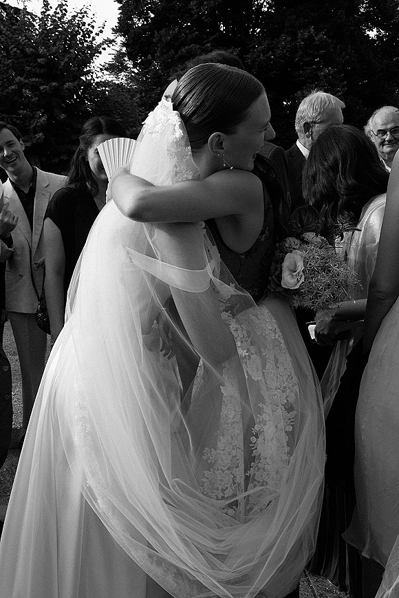 Wedding hug as bride embraces a guest, her long veil and lace dress visible amid outdoor trees and gathered wedding guests