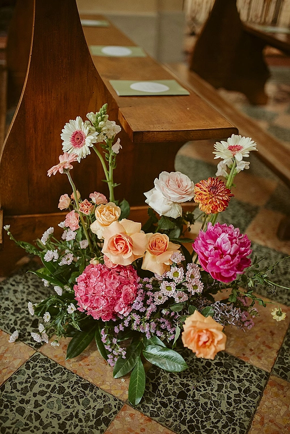 Ceremony aisle flowers in a grounded cluster of roses, peonies, hydrangea, and greenery beside wooden pews with programs