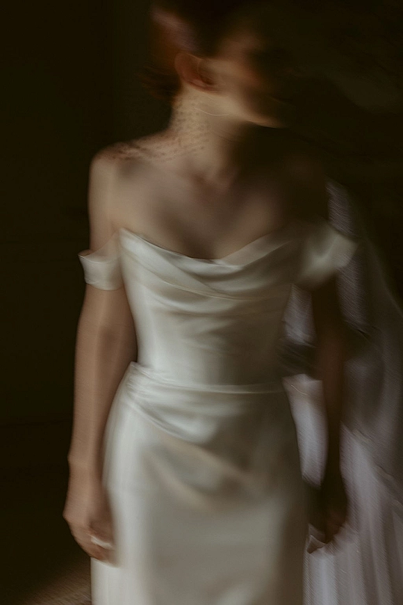 Bridal portrait in an off the shoulder wedding dress with satin drape, high neck lace top, and textured veil against a dark interior wall