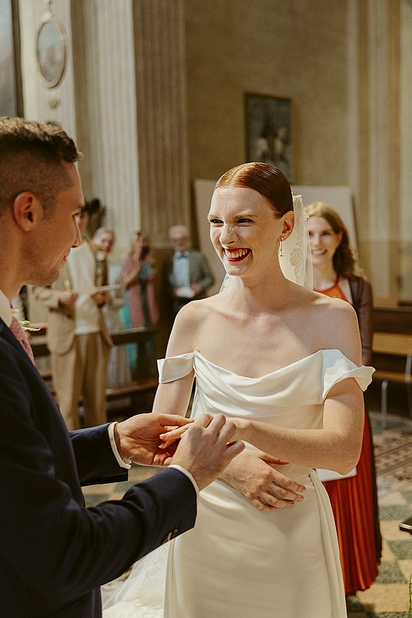 Wedding ceremony moment as groom places a wedding ring on the laughing bride’s hand, her veil trailing in a columned indoor space