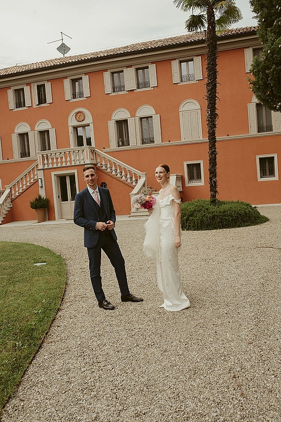 Couple portrait of bride and groom portrait, bride holding a colorful bouquet beside a European villa staircase in a gravel courtyard under clouds