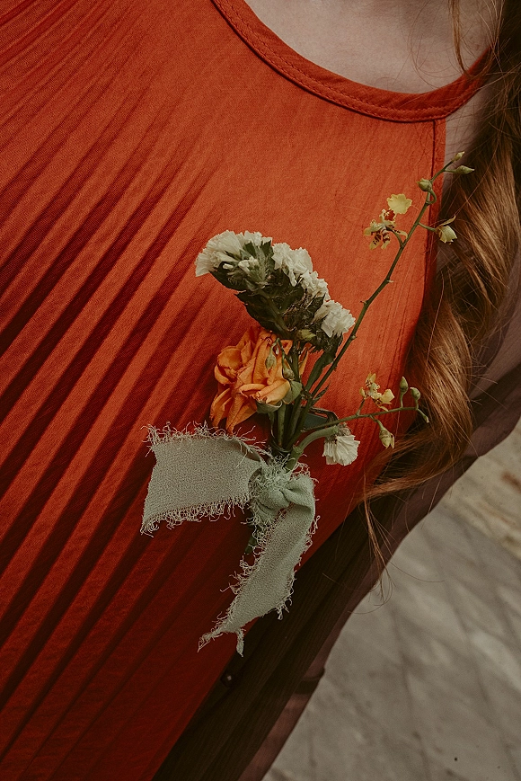 Boutonniere close-up of an orange rose boutonniere with white and yellow blooms and green ribbon bow pinned to a rust dress against sand ground