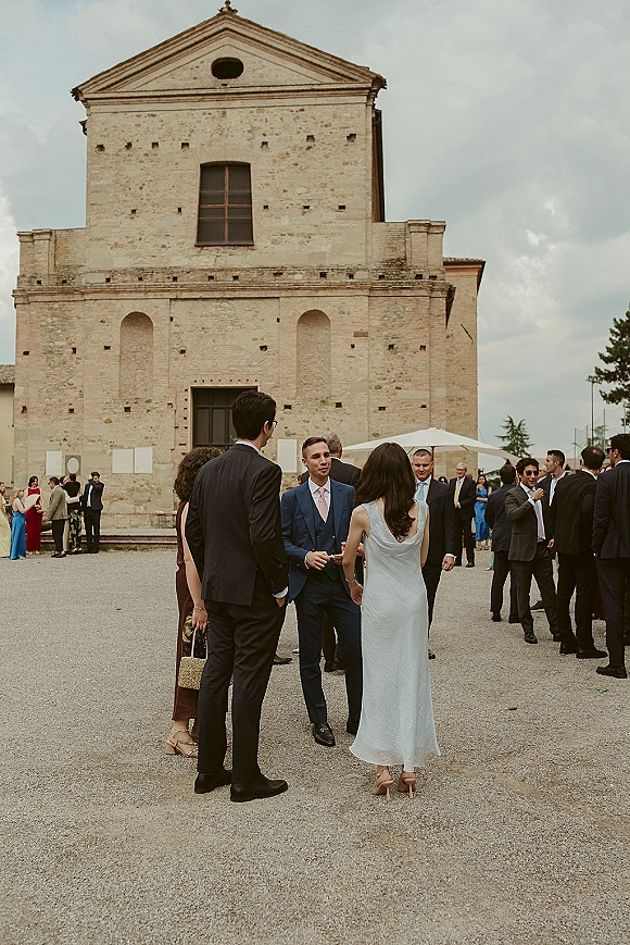 Wedding cocktail hour with wedding guests mingling in suits and cocktail dresses under patio umbrellas in a stone church courtyard