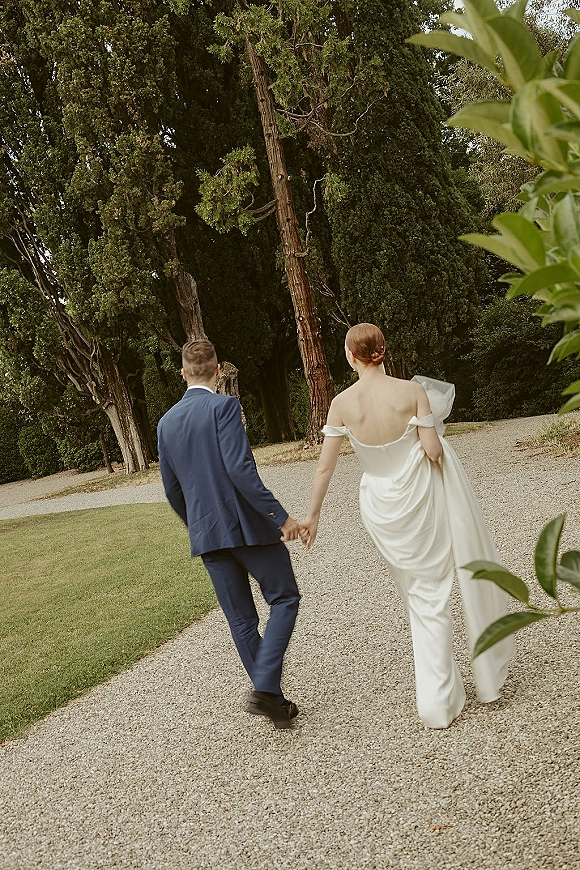 Couple portrait of bride and groom walking away holding hands, her veil blowing behind a simple satin dress on a gravel path in greenery