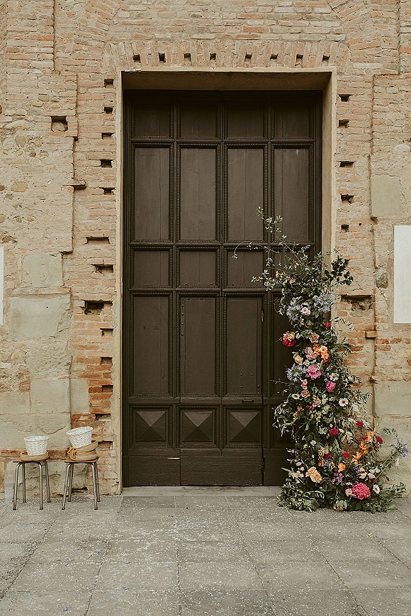 Wedding floral installation with ceremony entrance flowers framing a large wooden door, lush greenery and wicker baskets against brick and stone walls