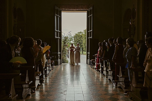 Wedding processional as bride walking down aisle with bouquet and veil, escorted by bridesmaid, guests with fans in backlit church doorway