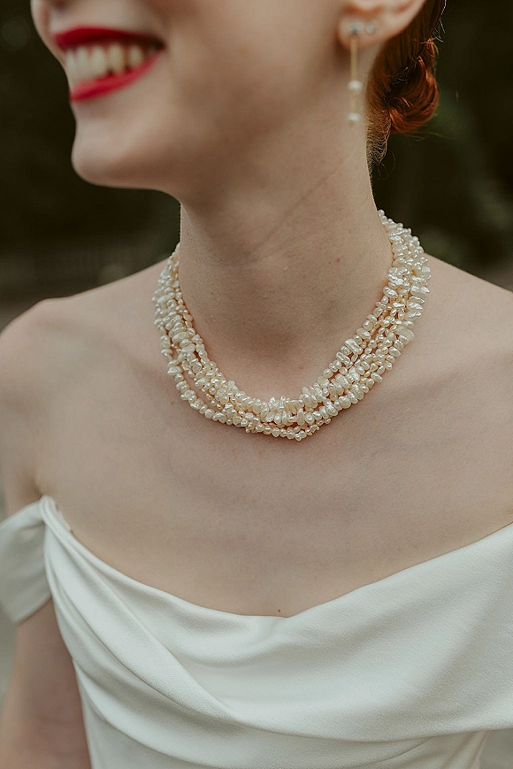 Bridal jewelry with a multi strand pearl necklace and pearl drop earrings on a bride in a strapless satin gown, blurred greenery behind