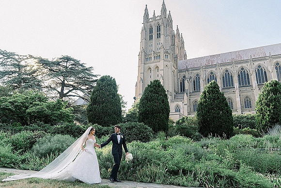 Couple portrait of bride and groom holding hands, her long veil blowing, by cathedral exterior with trees and stone walkway