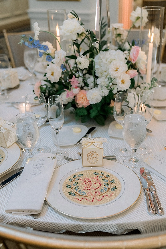 Reception tablescape with wedding table centerpiece of low florals and taper candles in hurricane vases, gold-rim plates and menu cards indoors
