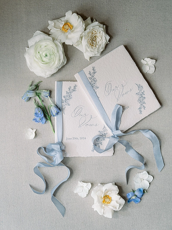 Wedding vow books tied with a silk ribbon, floral illustrations and blue-and-white petals arranged on a linen fabric backdrop