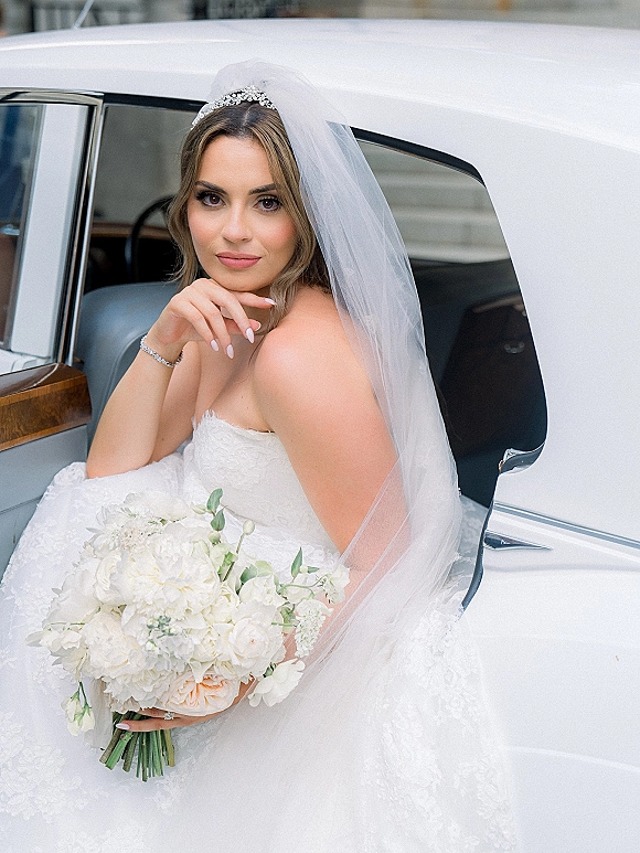 Bridal portrait of a bride in wedding car holding a white bouquet, wearing a tiara and veil, framed by the car interior and neutral building backdrop