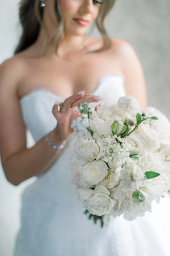 Bridal portrait of a bride holding bouquet of white roses and greenery in a strapless wedding dress, softly lit against a neutral wall
