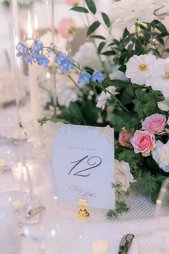 Wedding table number displayed in a gold holder beside roses, blue delphinium, and taper candles on a reception table with bokeh lights