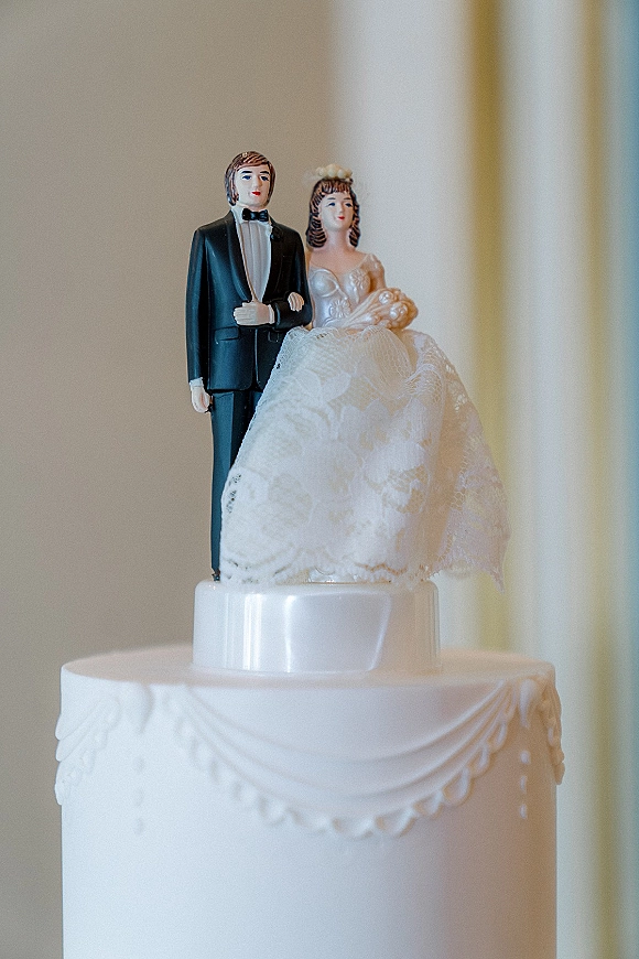 Wedding cake topper with a bride and groom figurine in tuxedo and wedding dress on a white cake, lace accent, soft indoor light