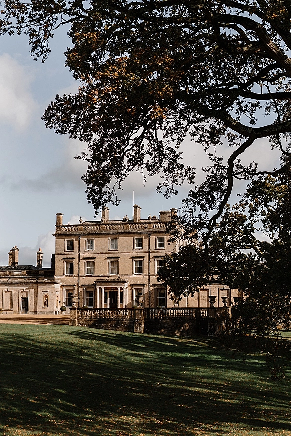 Wedding venue exterior of a historic manor house with neoclassical columns and a stone balustrade overlooking a wide lawn and trees under clouds