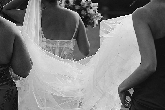 Bridal veil moment as bridesmaids fixing veil on bride from behind, lace spaghetti-strap gown and bouquet in blurred greenery outdoors