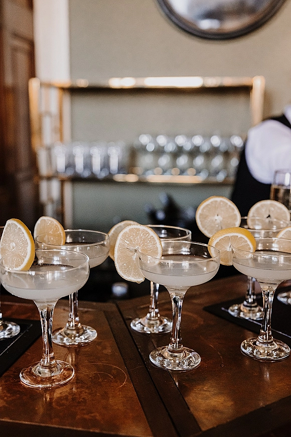 Wedding cocktails in coupe glasses with lemon garnish and cocktail napkins on a wooden bar top, with mirrored bar shelves behind