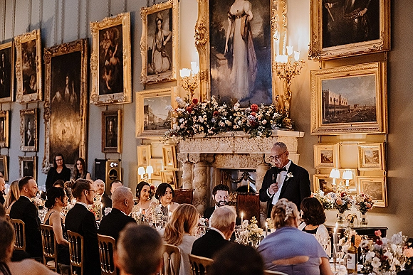 Wedding speech with father of the bride speech in a tuxedo holding a microphone, candles and florals glowing by a fireplace in a gallery-walled hall