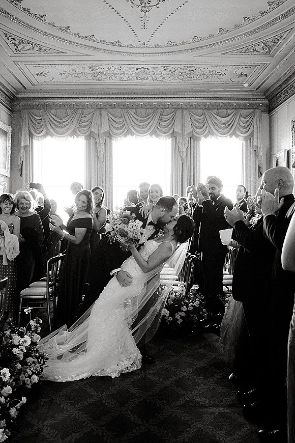 Wedding kiss portrait of newlyweds sharing an aisle dip kiss, bride in lace dress and veil with bouquet as guests cheer by tall windows
