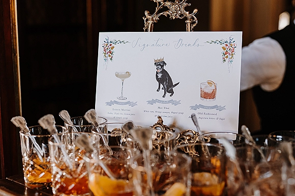 Signature drinks sign in a gold ornate frame on a bar cart, with cocktail glasses, stir sticks, lemon twist and ice in a dark wooden interior