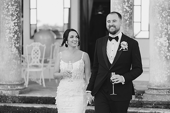 Newlywed couple portrait with bride and groom holding hands, raising champagne flutes on venue steps by stone columns and windows