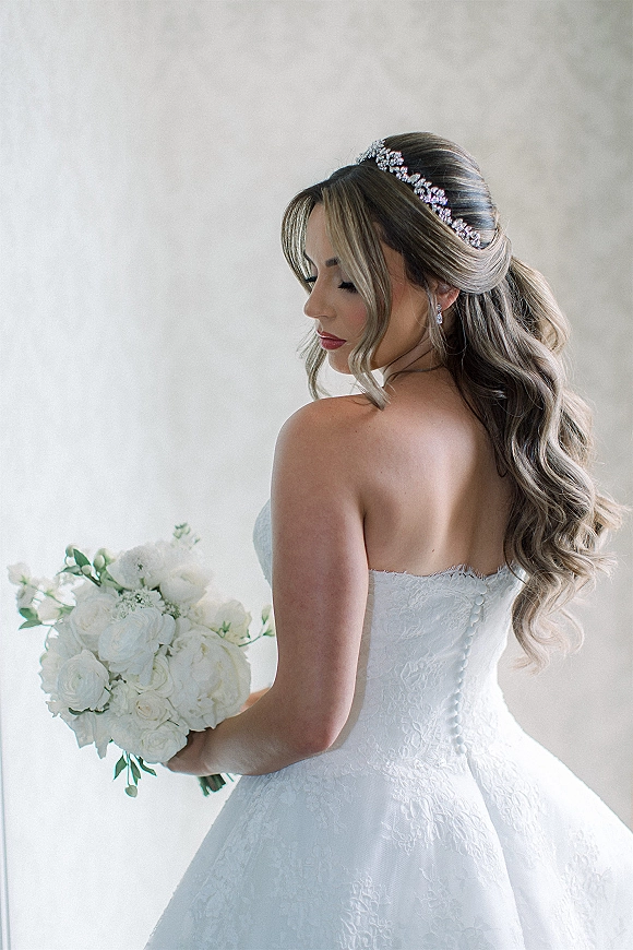 Bridal portrait of a bride holding bouquet of white roses and greenery, wearing a strapless lace dress and crystal headband indoors