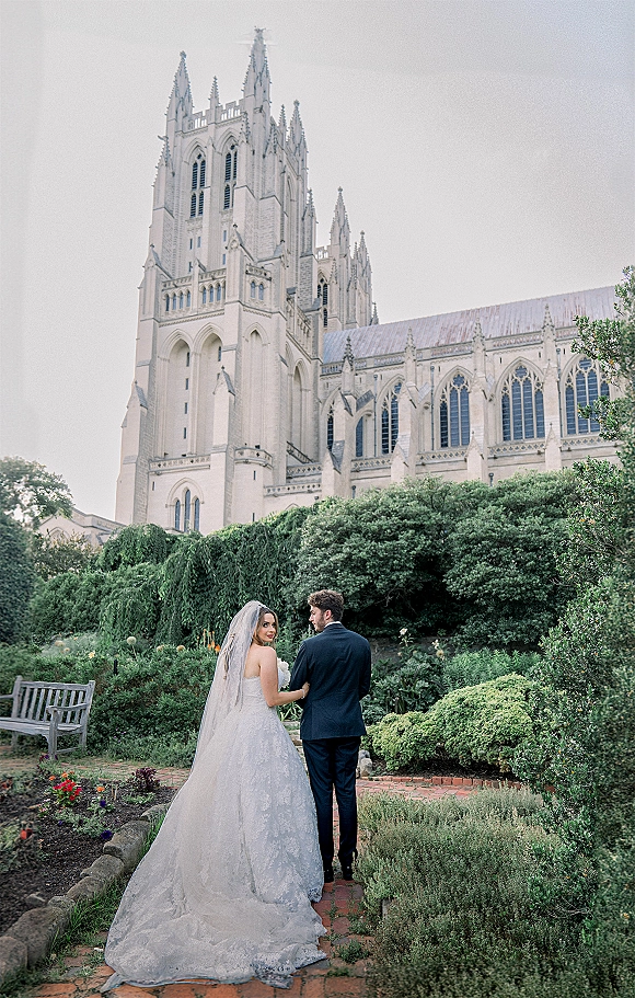 Couple portrait of bride and groom walking away on a brick path, bride looking back as her veil and lace train trail by a cathedral garden