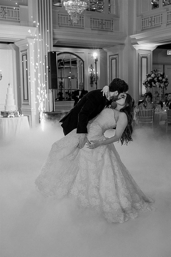 First dance kiss as groom dips bride in gown amid sparkler fountains and low fog, beneath chandelier in grand ballroom with guests seated