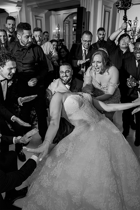 Wedding dance moment as bride in a strapless lace ballgown is lifted by guests in a ballroom, while a videographer films on a gimbal