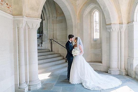 Couple portrait of bride and groom embracing under stone arches, her veil and long train flowing as she holds a white bouquet