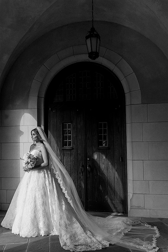 Bridal portrait of a bride holding bouquet in a strapless lace wedding gown with long lace veil, standing in a sunlit stone archway doorway