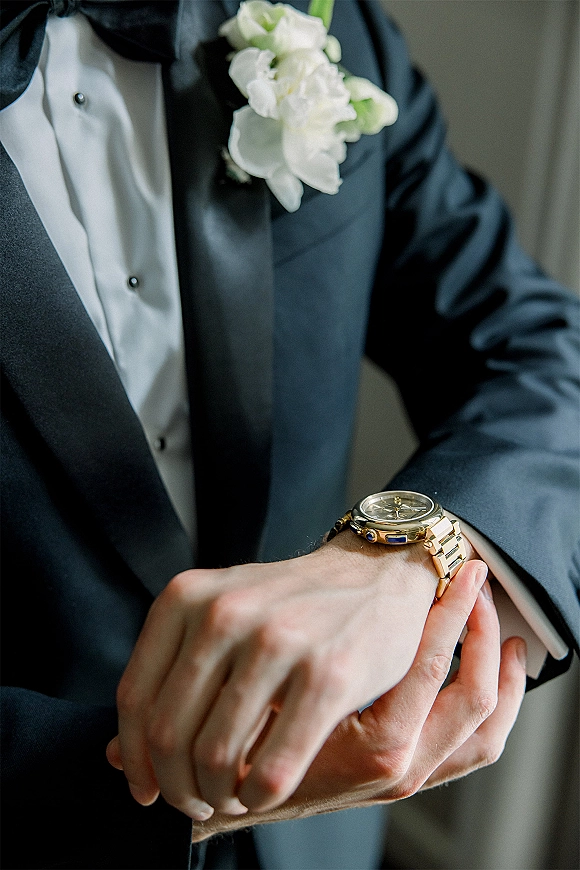 Groom getting ready adjusts his wristwatch over a crisp white cuff, black tuxedo and bow tie with a white boutonniere against a neutral wall