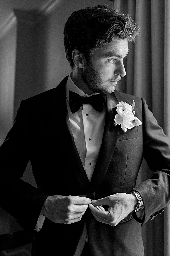 Groom portrait in a black and white wedding portrait style, wearing tuxedo with bow tie and boutonniere by a window with curtains indoors