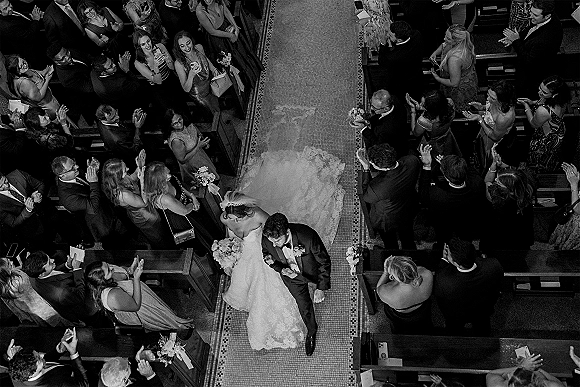 Wedding recessional overhead wedding photo of bride and groom walking church aisle, her long train and veil flowing as guests cheer from pews