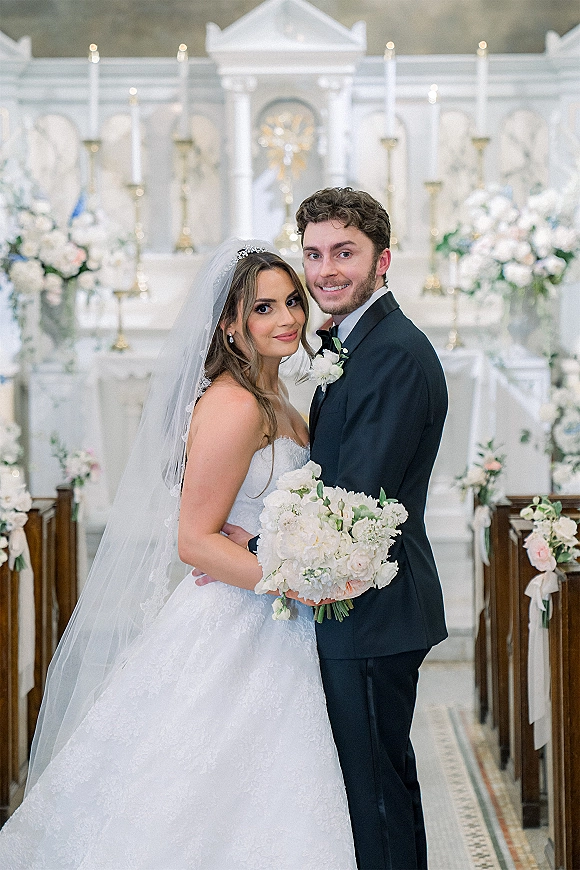 Couple portrait of bride and groom holding a bouquet in a church wedding portrait, with veil, tuxedo, and candles at the altar