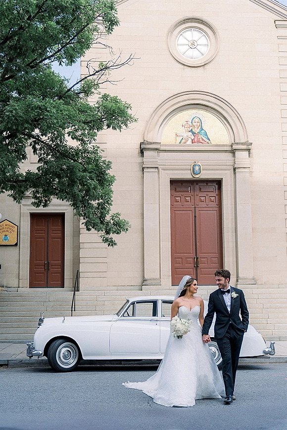 Couple portrait of bride and groom walking hand in hand, her veil and bouquet flowing beside a white vintage car at church steps