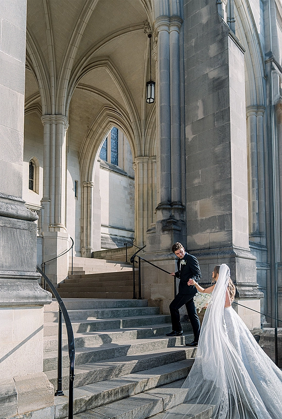 Couple portrait of bride and groom on steps, holding hands as he helps her, her veil and bouquet flowing by a stone church archway