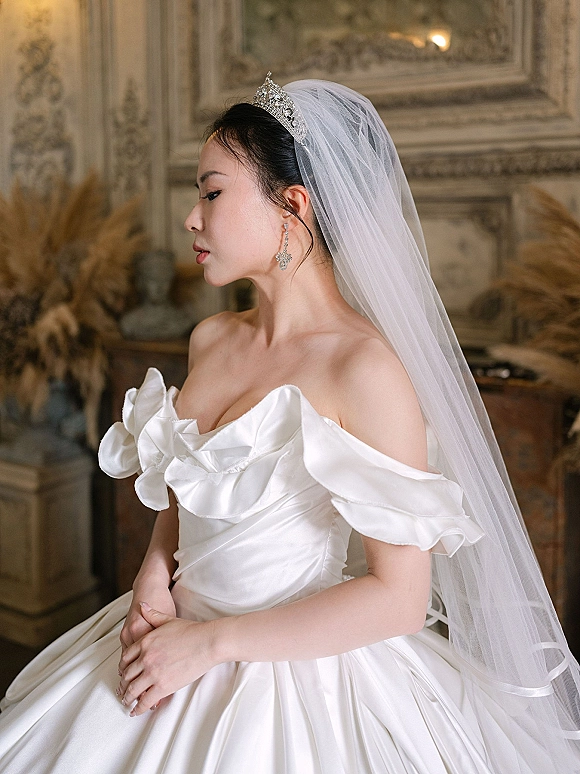 Bridal portrait of a side profile bride in a tiara and veil, wearing a satin off-shoulder gown before ornate wall paneling and art
