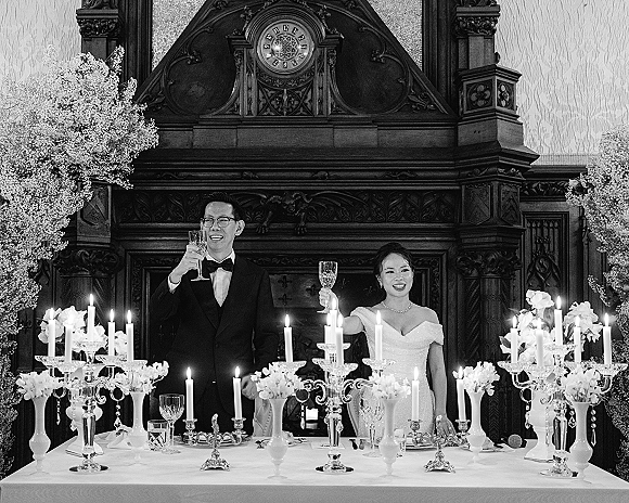 Wedding toast as bride and groom raise champagne glasses at a candlelit sweetheart table with candelabras before an ornate mantel clock