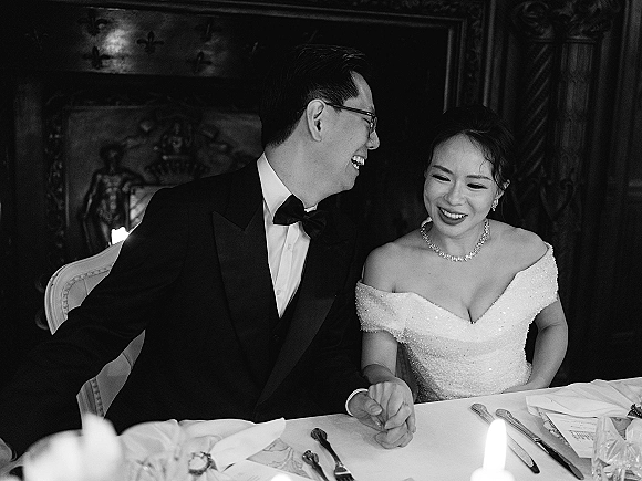 Wedding couple portrait in a black and white wedding portrait, bride in off shoulder gown and groom in tuxedo laughing at a candlelit table
