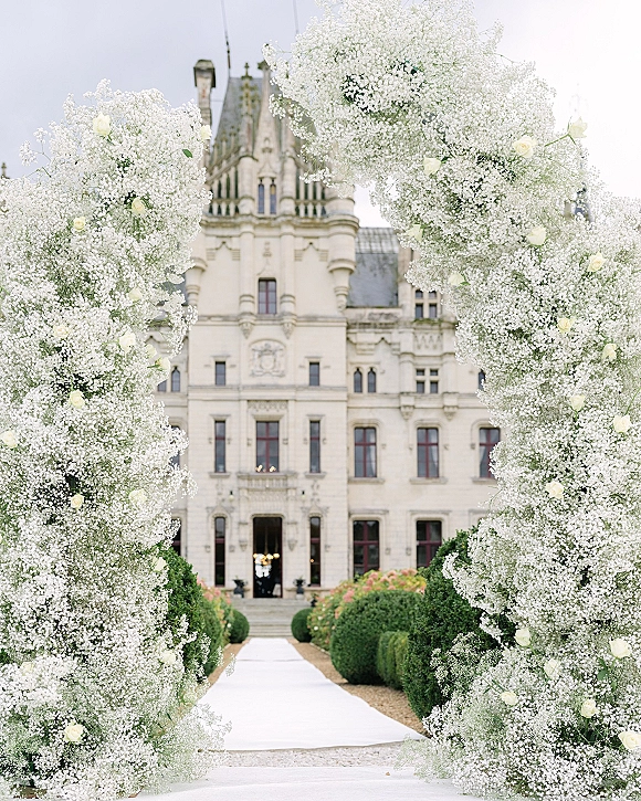 Ceremony aisle decor with baby's breath and white roses flanking a white runner on a gravel path toward a castle facade under open sky