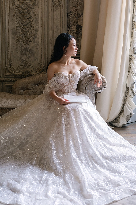 Bridal portrait of a bride by window in a strapless lace gown with long train, seated on a vintage sofa holding a paper letter