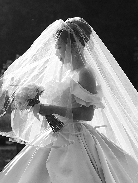 Bridal portrait in black and white of a bride holding bouquet, looking down beneath a flowing veil, with outdoor greenery behind her