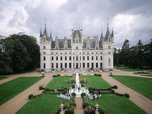 Outdoor wedding ceremony with a white aisle runner flanked by floral arrangements and chairs set before a castle facade in a formal garden
